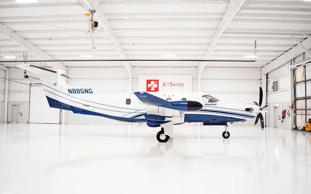 A white and blue Pilatus PC-12 aircraft, tail number N885NG, is parked in a brightly lit hangar, viewed from the side.