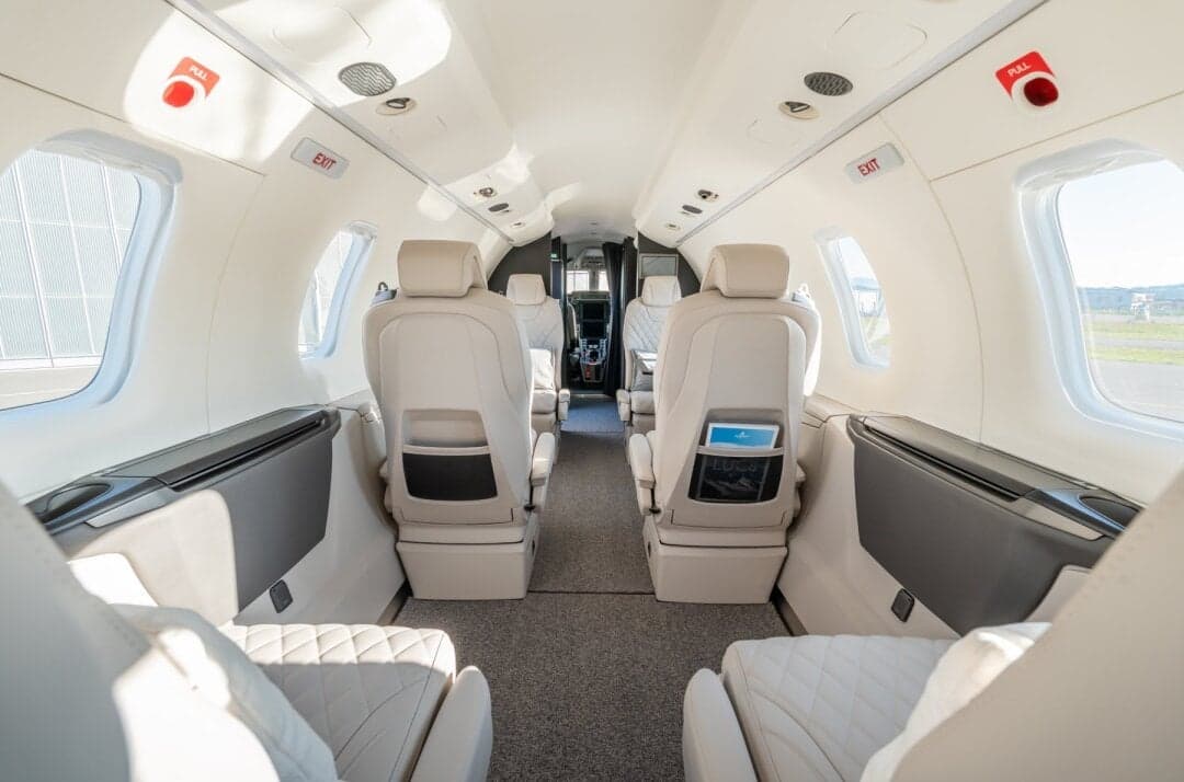 A view down the aisle of a private jet cabin, showing light-colored quilted leather seats and large windows.