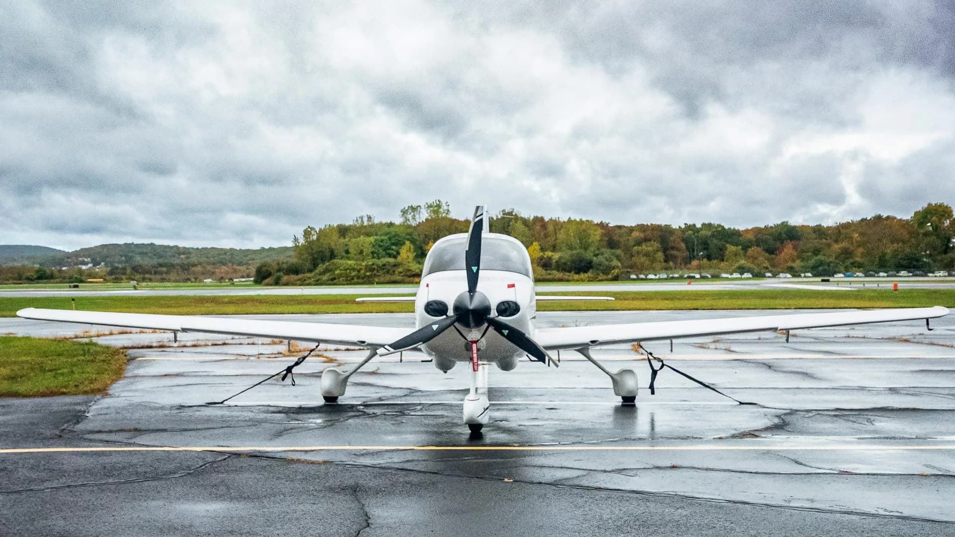 A head-on view of a white Cirrus SR20 aircraft with registration N899WT, parked on a wet tarmac under a cloudy sky.