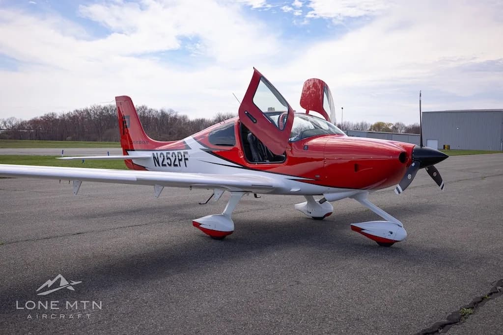 A front-side view of a red and white Cirrus SR20 aircraft with its doors open, parked on an airfield.