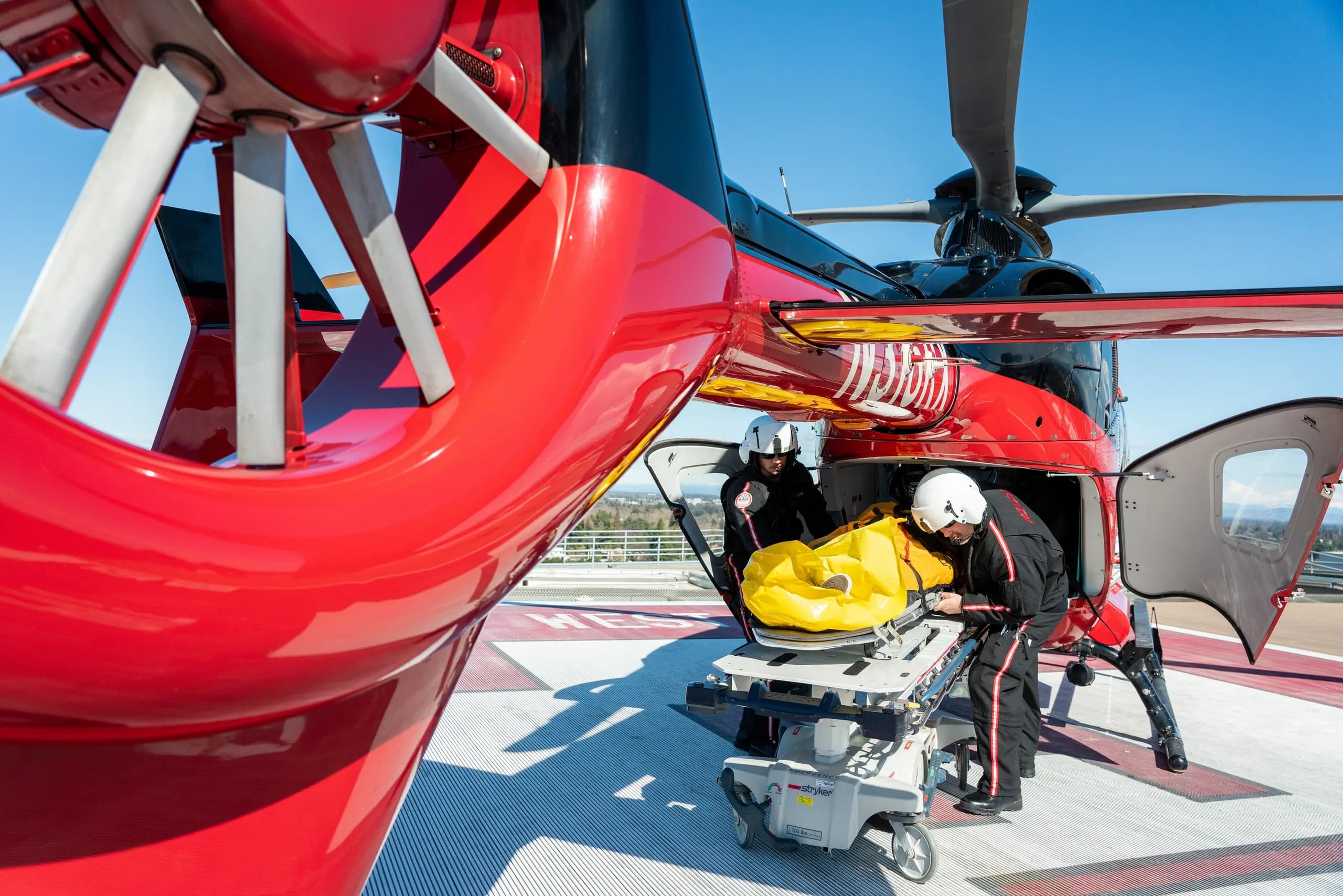 A red and black Airbus EC135 helicopter with medical personnel loading a stretcher.