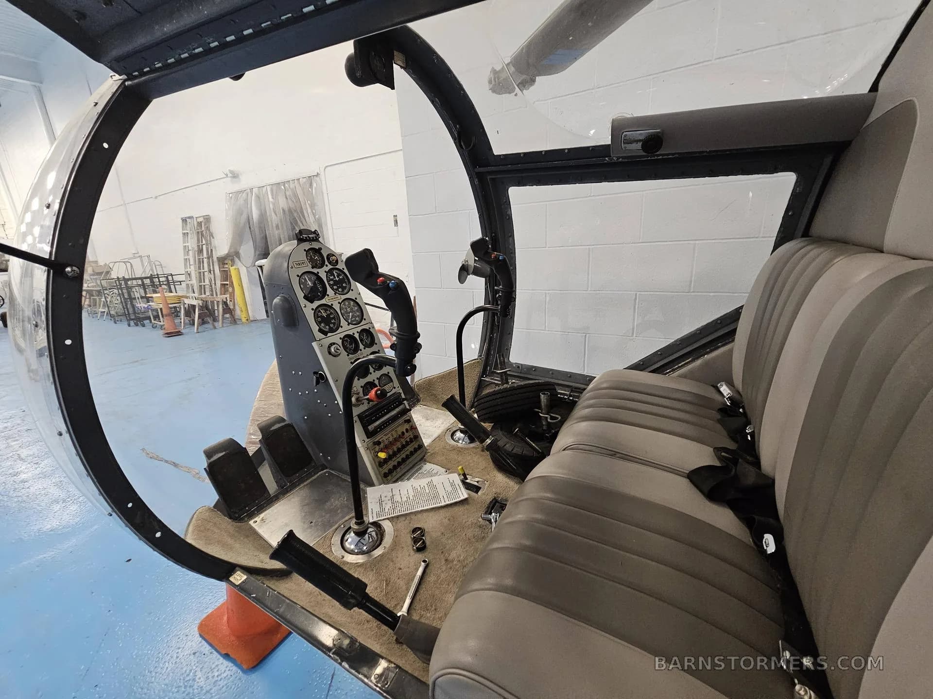 The cockpit of a helicopter with a grey instrument panel, controls, and two-tone grey seats, viewed through the clear canopy.
