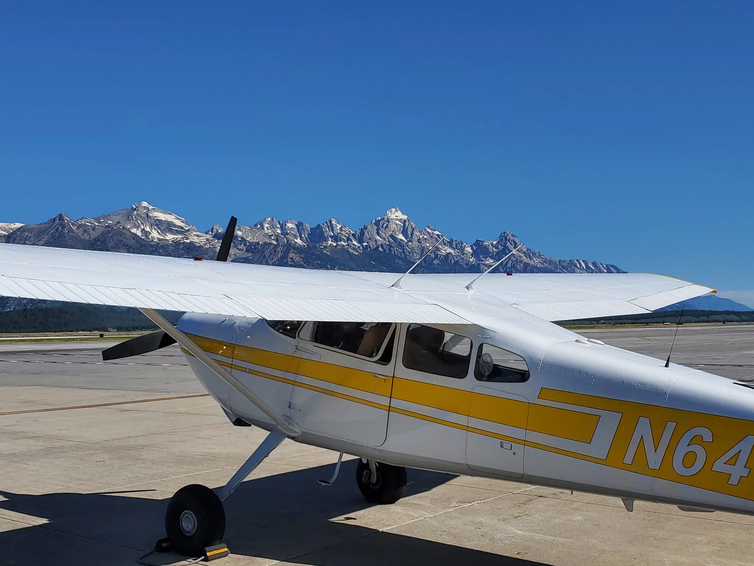 A white and yellow Cessna 172 aircraft with registration N644FM parked on an airfield with snow-capped mountains in the background.