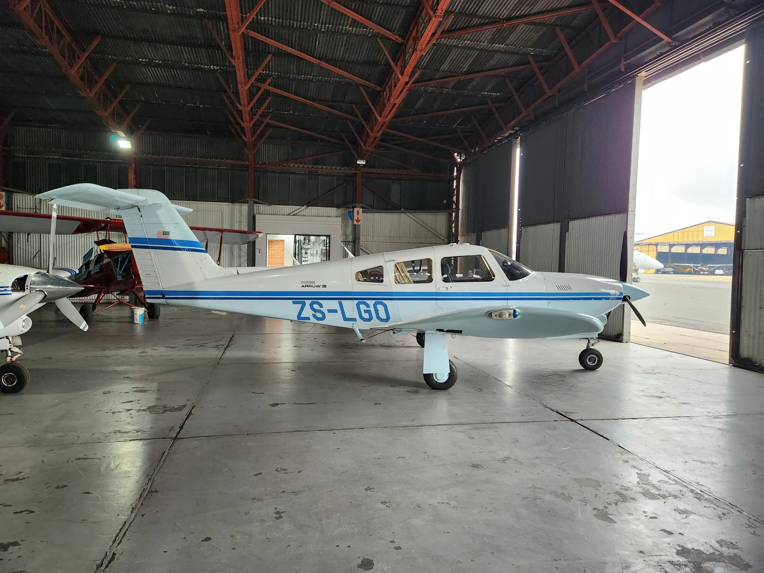 A white and blue Piper aircraft, registration ZS-LGO, is parked inside a hangar with the door open, viewed from the right side.