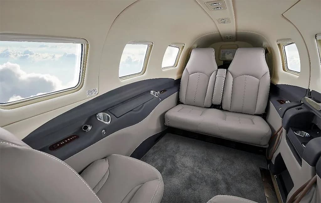 The light gray and dark gray interior cabin of a small aircraft with two rows of seats and windows showing clouds.