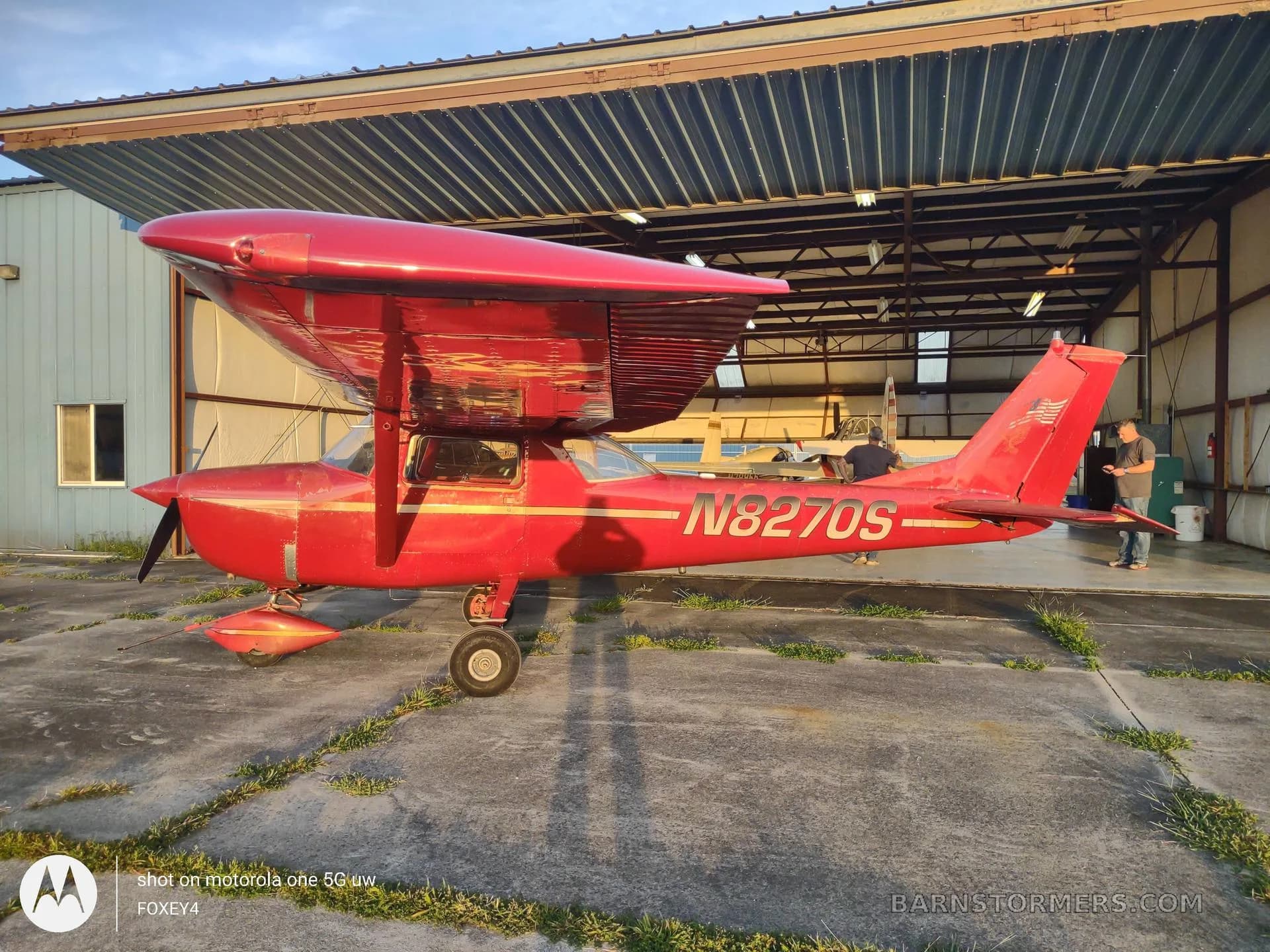 A side view of a red Cessna 150 or similar light aircraft with registration N8270S parked on a concrete apron outside a hangar.