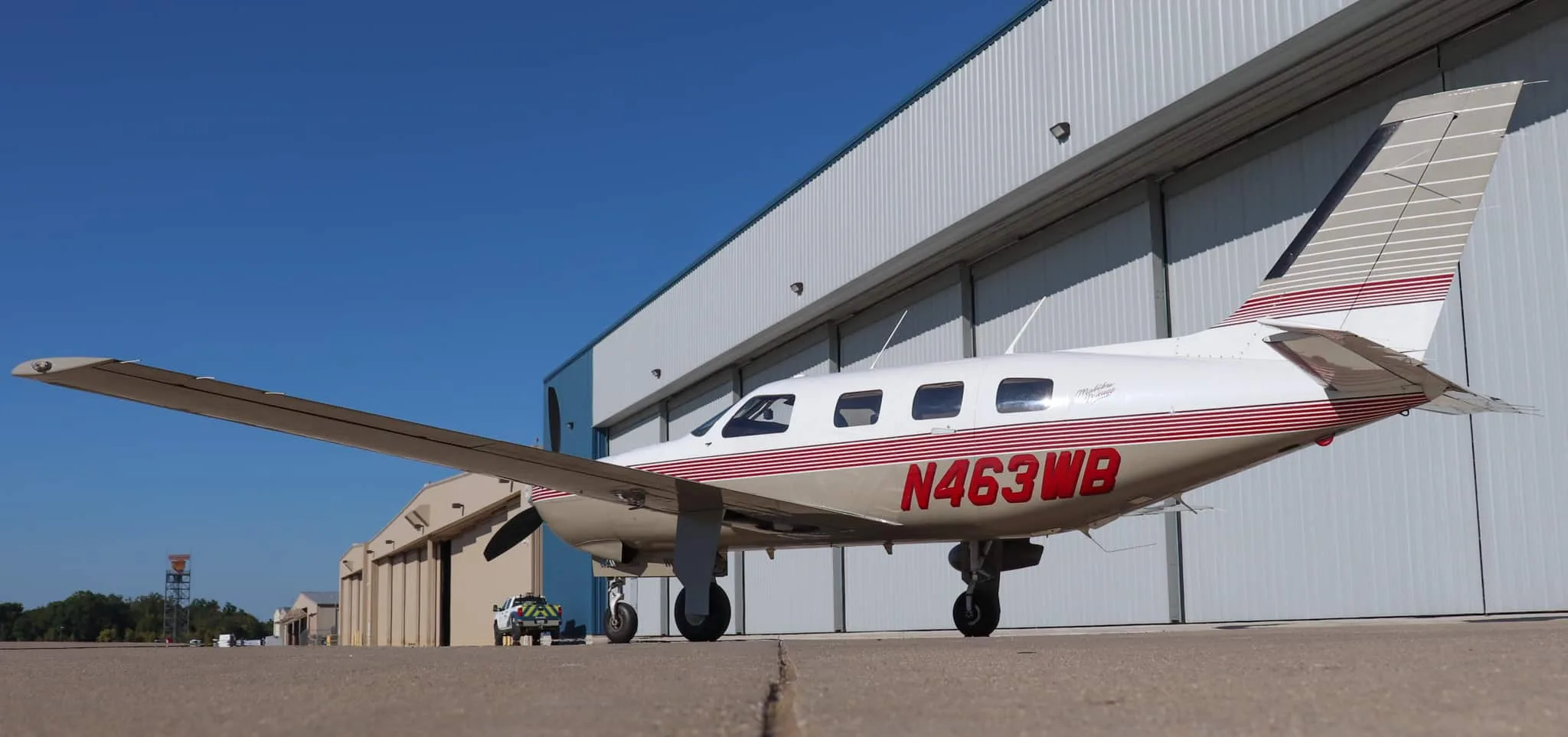 A low-angle side view of the Piper Malibu Mirage N463WB parked on an airport tarmac next to a large hangar.