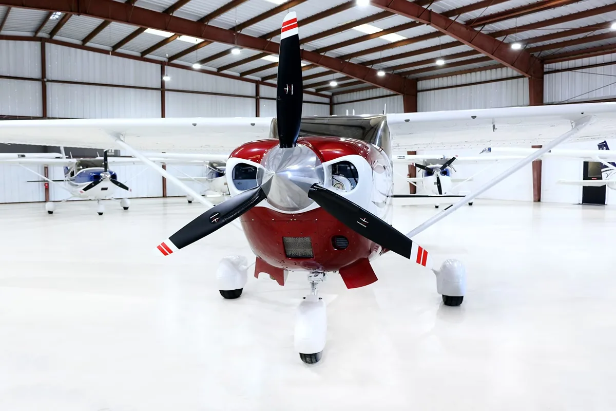 A head-on view of a red and white Cessna Stationair TC aircraft with a three-bladed propeller, parked in a hangar.
