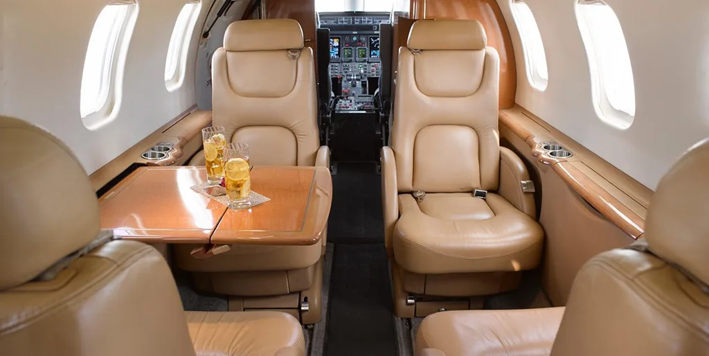 The aircraft cabin features tan leather seats and a wooden table with drinks, with the cockpit visible in the background.
