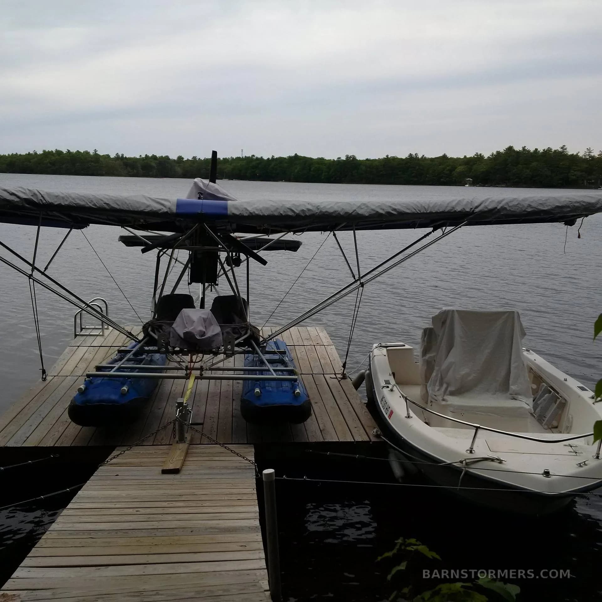 A blue ultralight seaplane with a gray covered wing docked on a wooden pier next to a white boat on a lake.