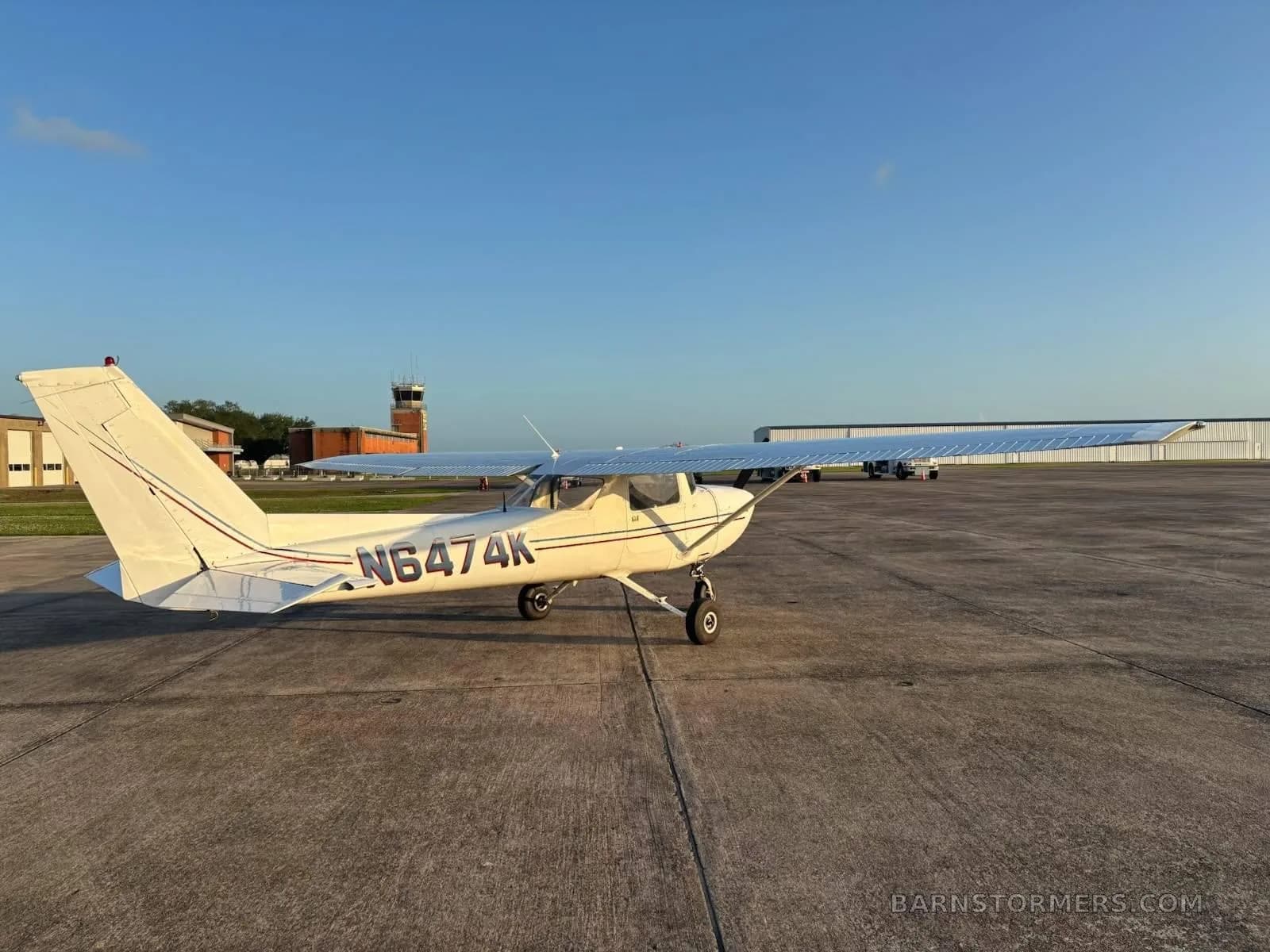 A white Cessna 172 (or similar) with tail number N6474K is parked on an airfield tarmac with a control tower in the background.