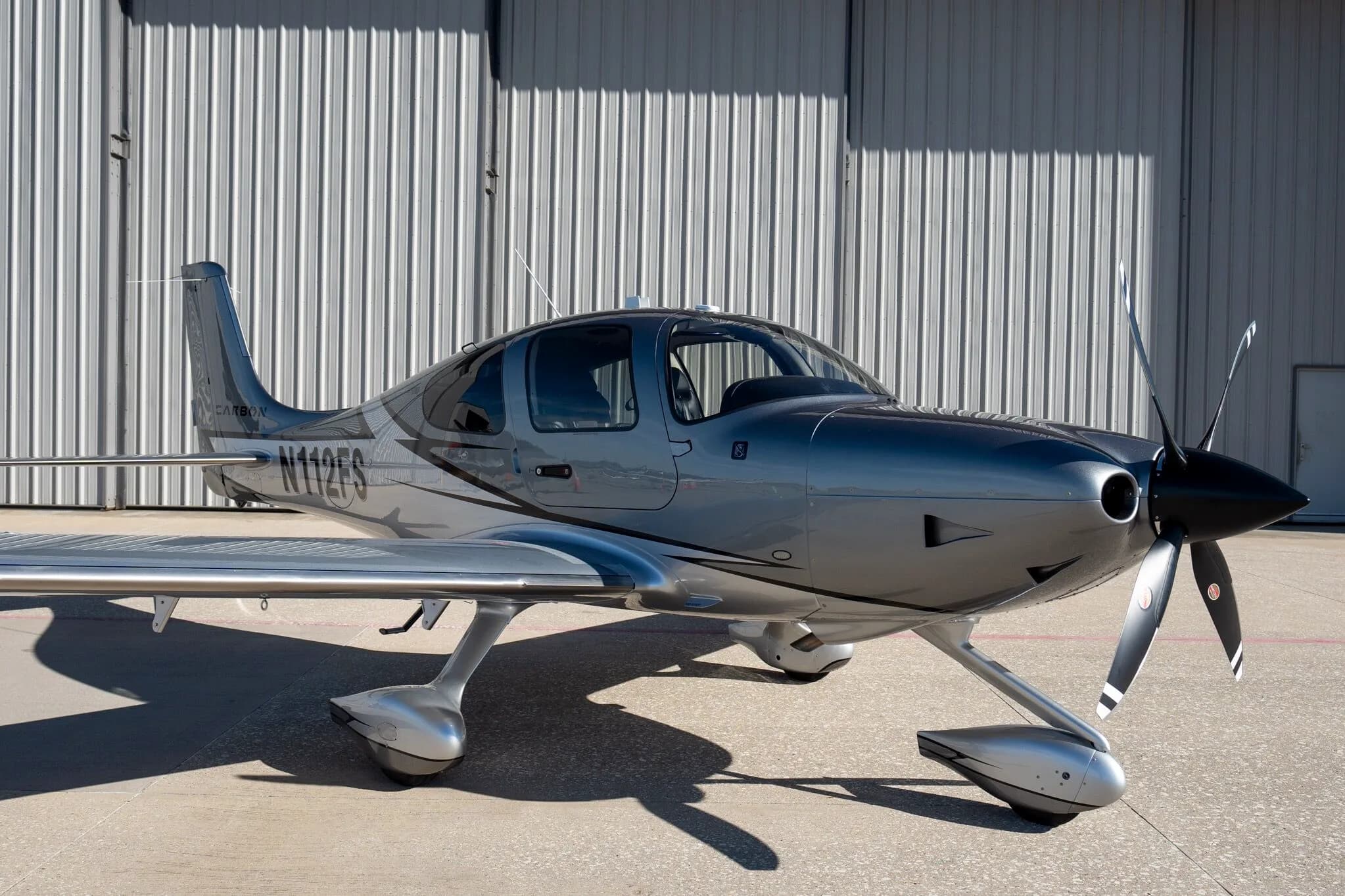 A front-right view of a silver and dark grey Cirrus SR22T Carbon Edition aircraft (N112FS) with a hangar in the background.