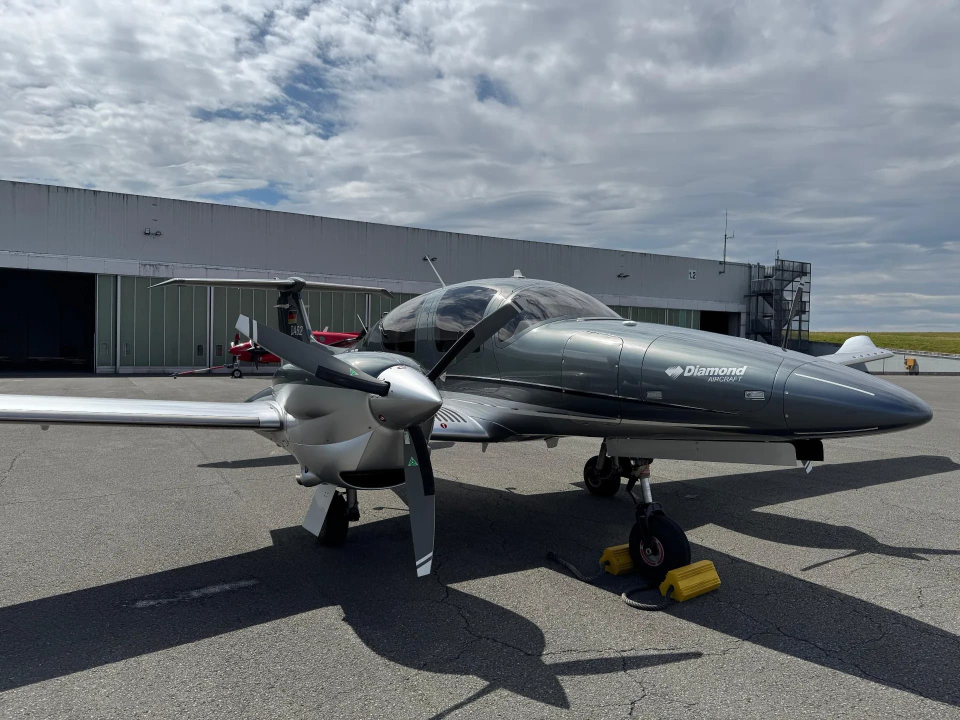 A dark grey Diamond DA62 twin-engine aircraft with silver accents parked on an asphalt tarmac in front of a hangar under a partly cloudy sky.