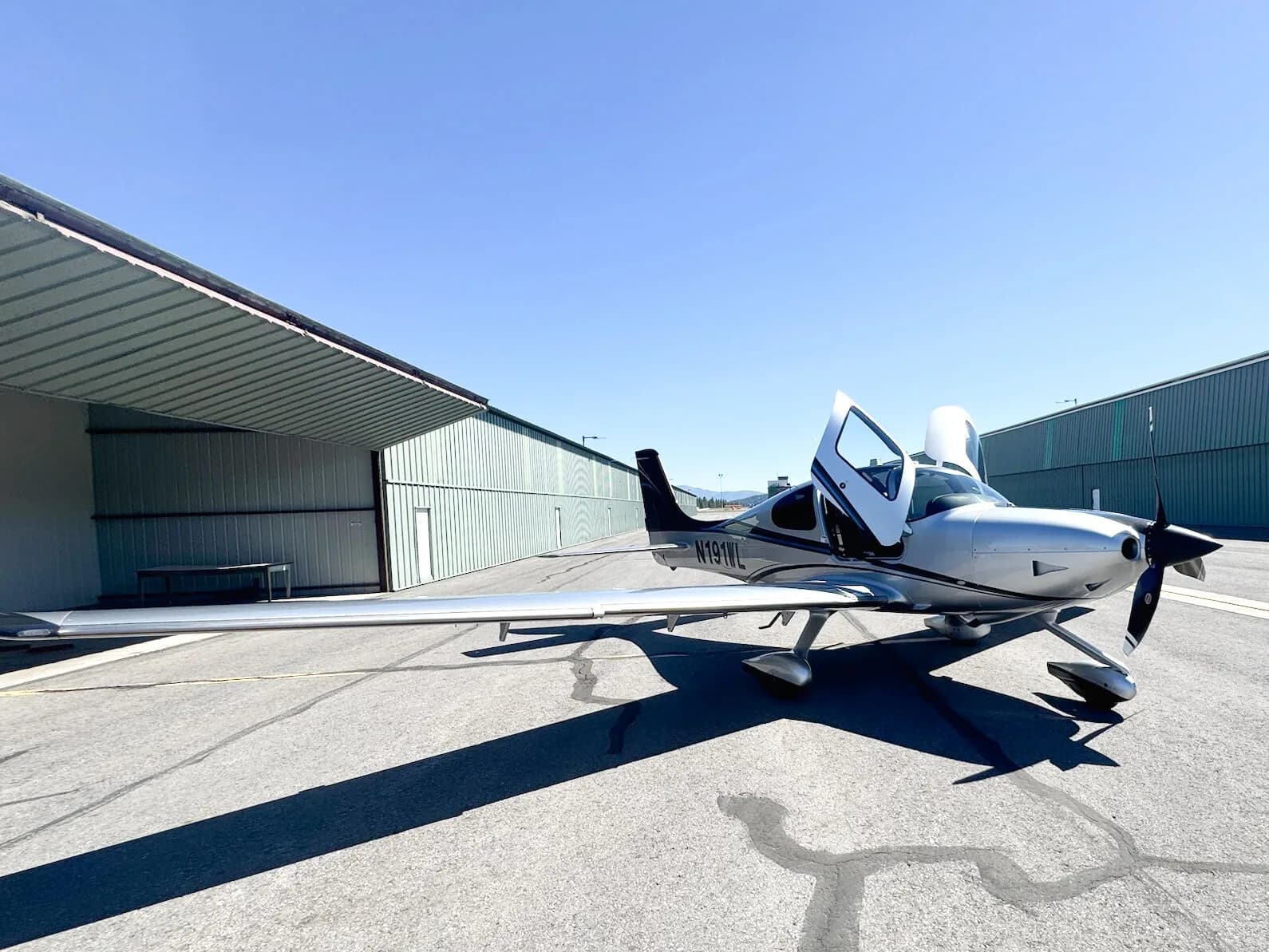 A wide-angle view of a silver and black Cirrus SR22T aircraft with N191WL, with both doors open, parked on an apron between hangars.