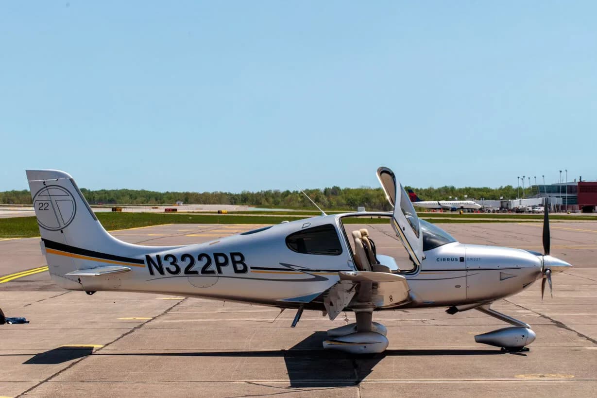 A silver Cirrus SR22T aircraft with registration N322PB, parked on an airfield with its door open.