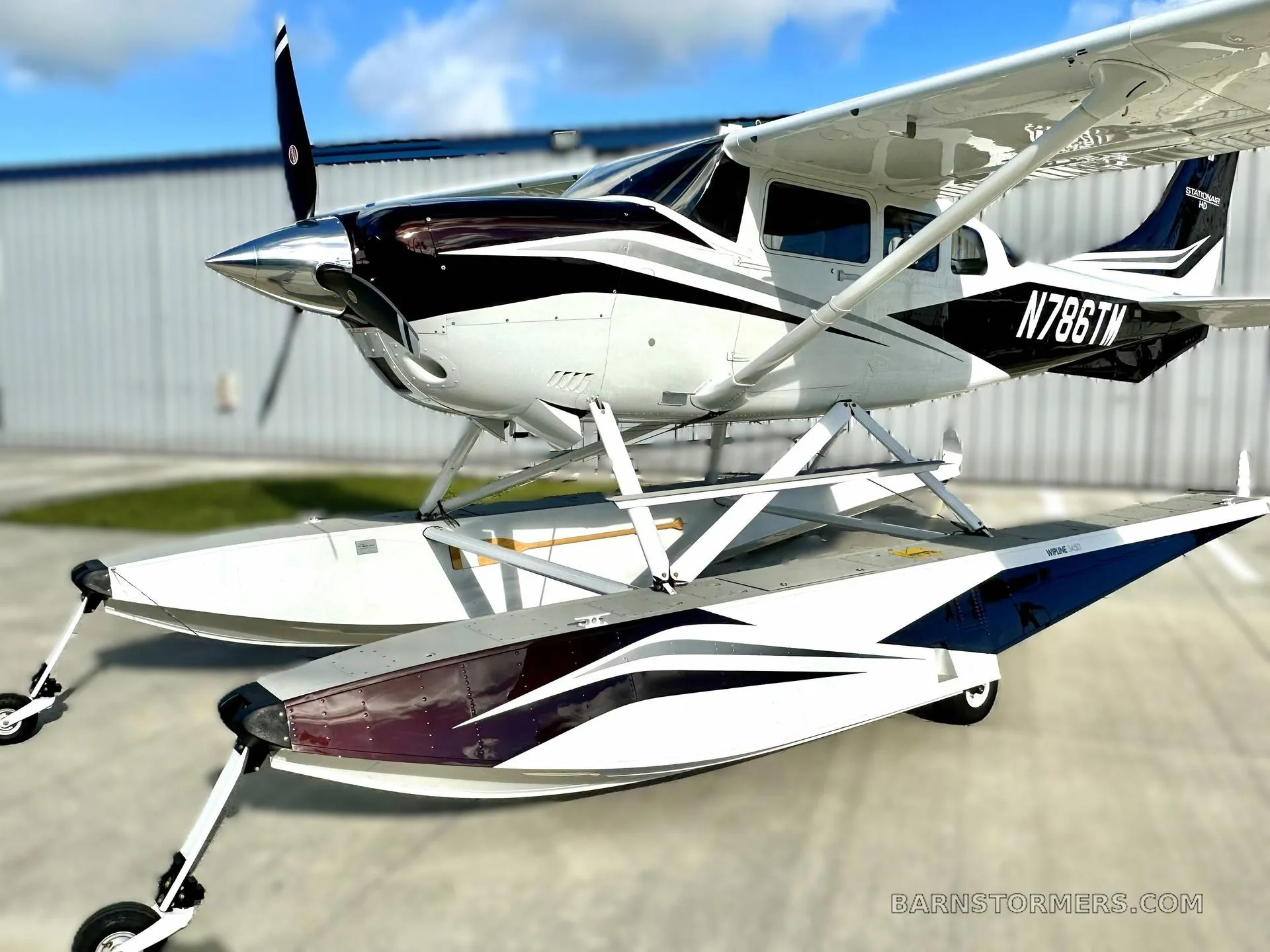 A white and black Cessna T206 floatplane with registration N786TM is parked on a tarmac under a blue sky.