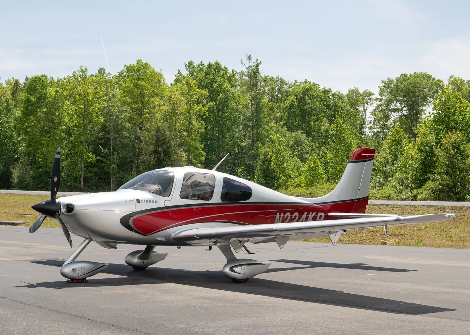 A silver and red Cirrus SR22 aircraft, N224KR, parked on an asphalt ramp with green trees in the background.