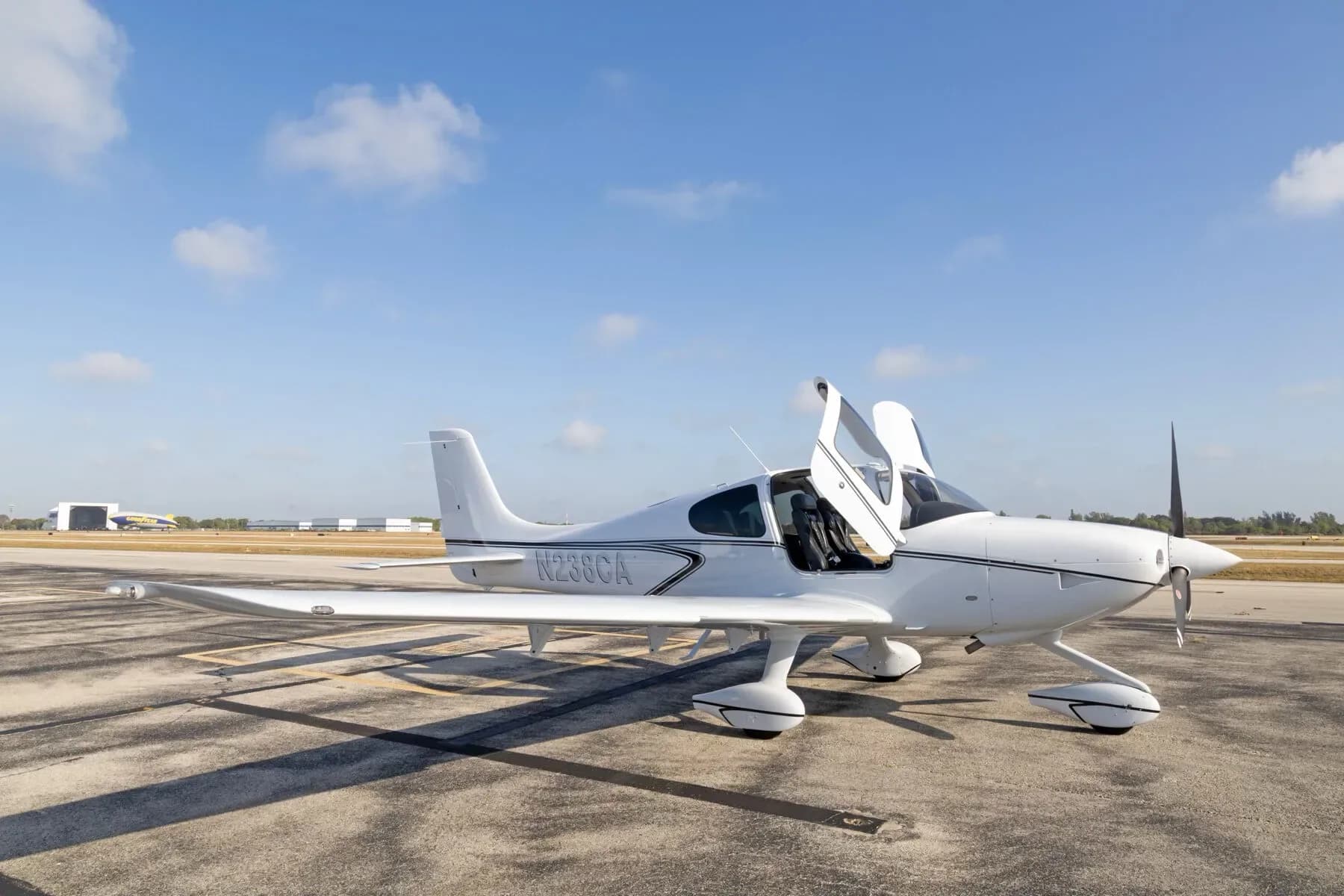 A white Cirrus SR22 aircraft with its gull-wing doors open is parked on an airport tarmac.