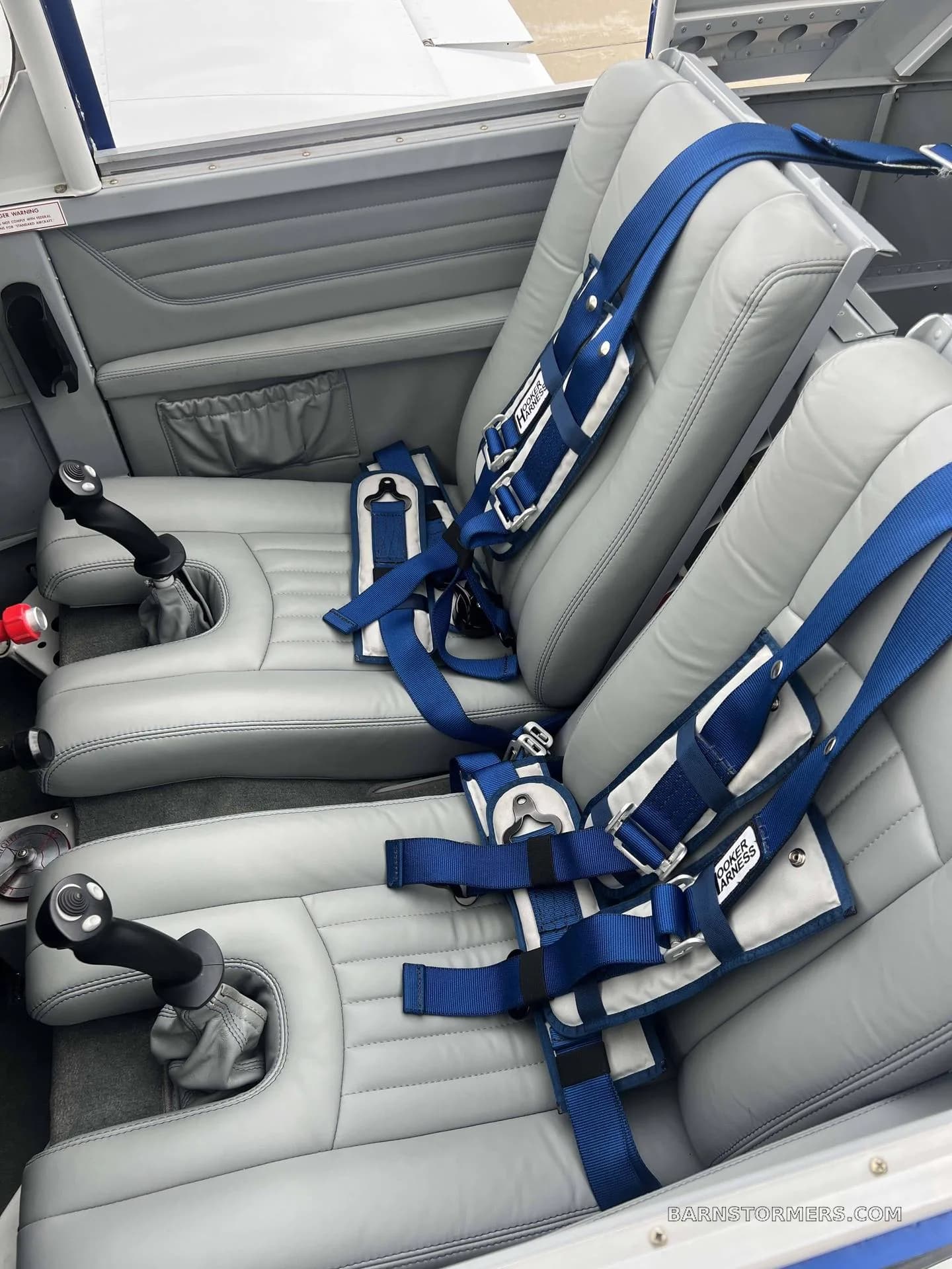 The interior cabin of a light aircraft, showing two grey leather seats with blue "HOOKER HARNESS" seatbelts and dual stick controls.