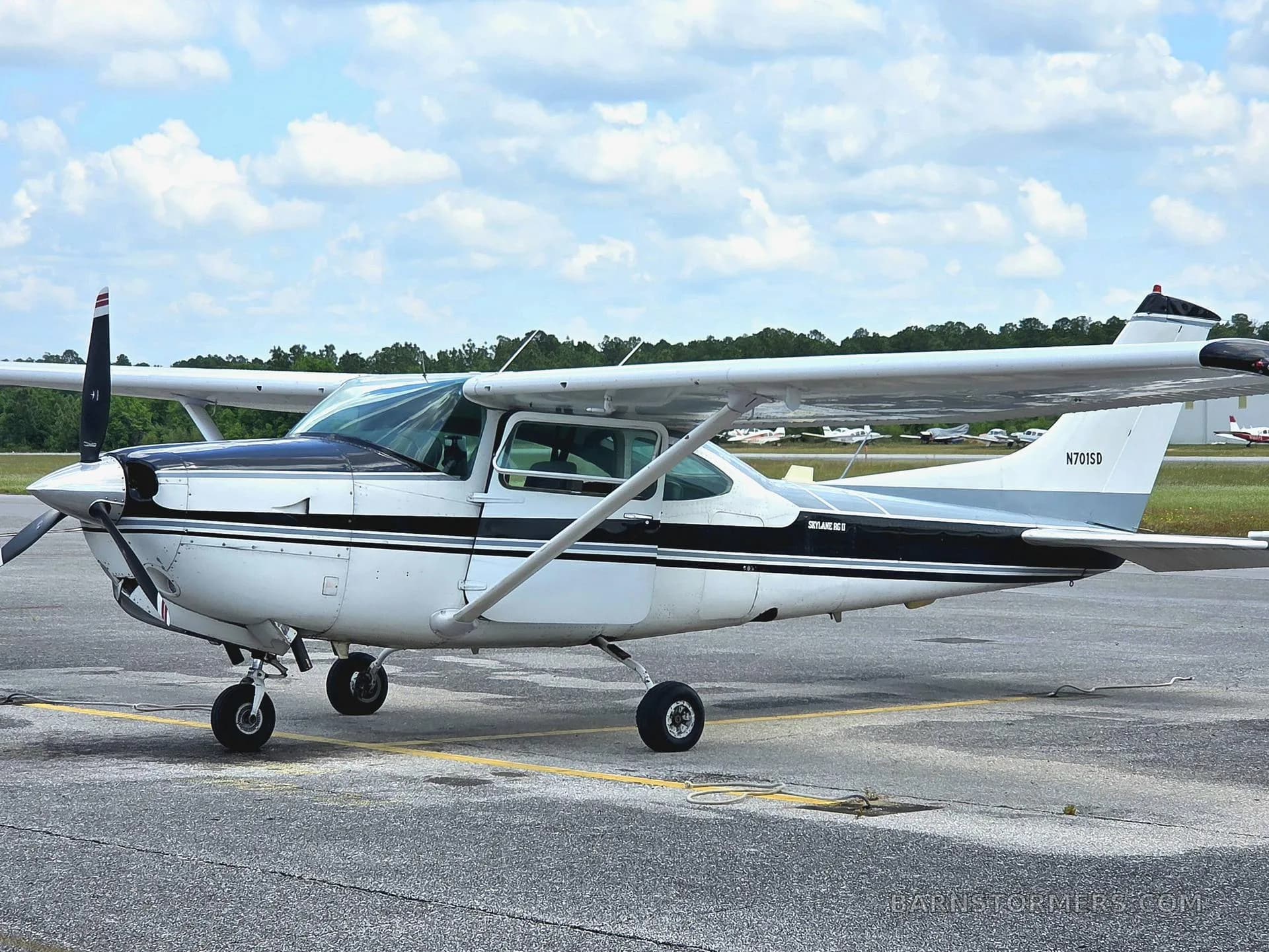 A white and black Cessna 182 Skylane with registration N701SD parked on an asphalt ramp under a partly cloudy sky.