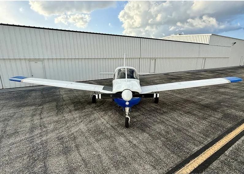 A head-on view of a white and blue Piper Arrow aircraft with registration N338PA parked on an asphalt tarmac in front of a hangar.