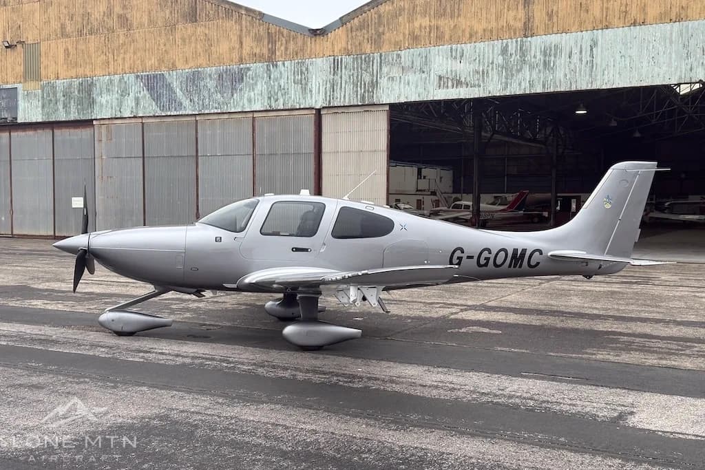 A side view of the silver Cirrus SR22 aircraft with its doors closed, parked on an airfield in front of a hangar.
