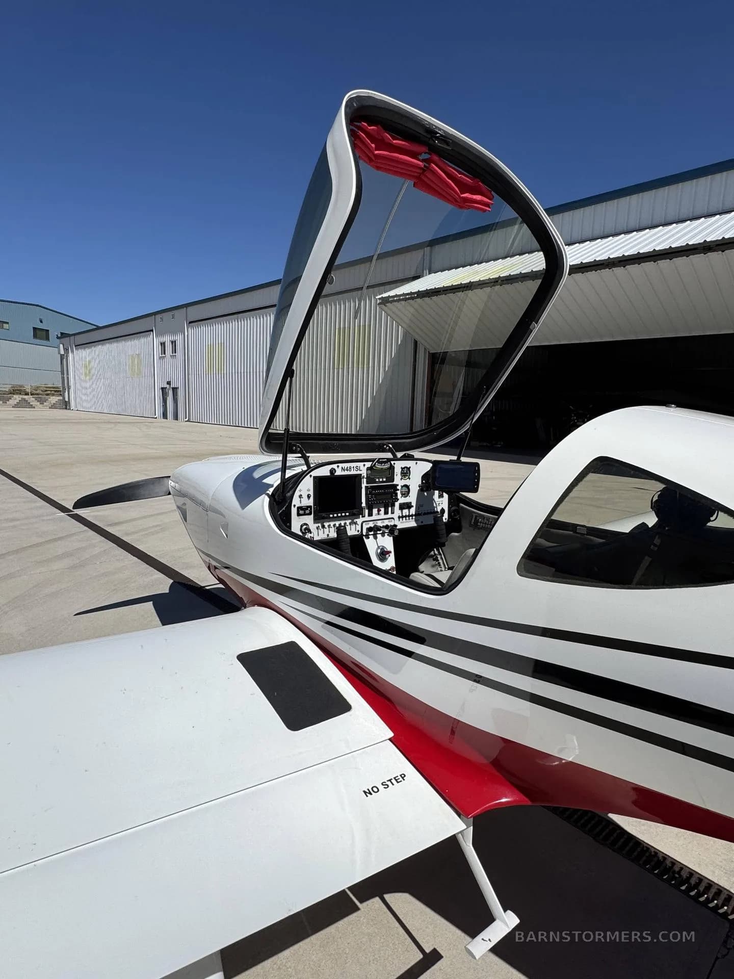 A white and red single-engine aircraft with its canopy open, revealing the cockpit, parked on an airfield.