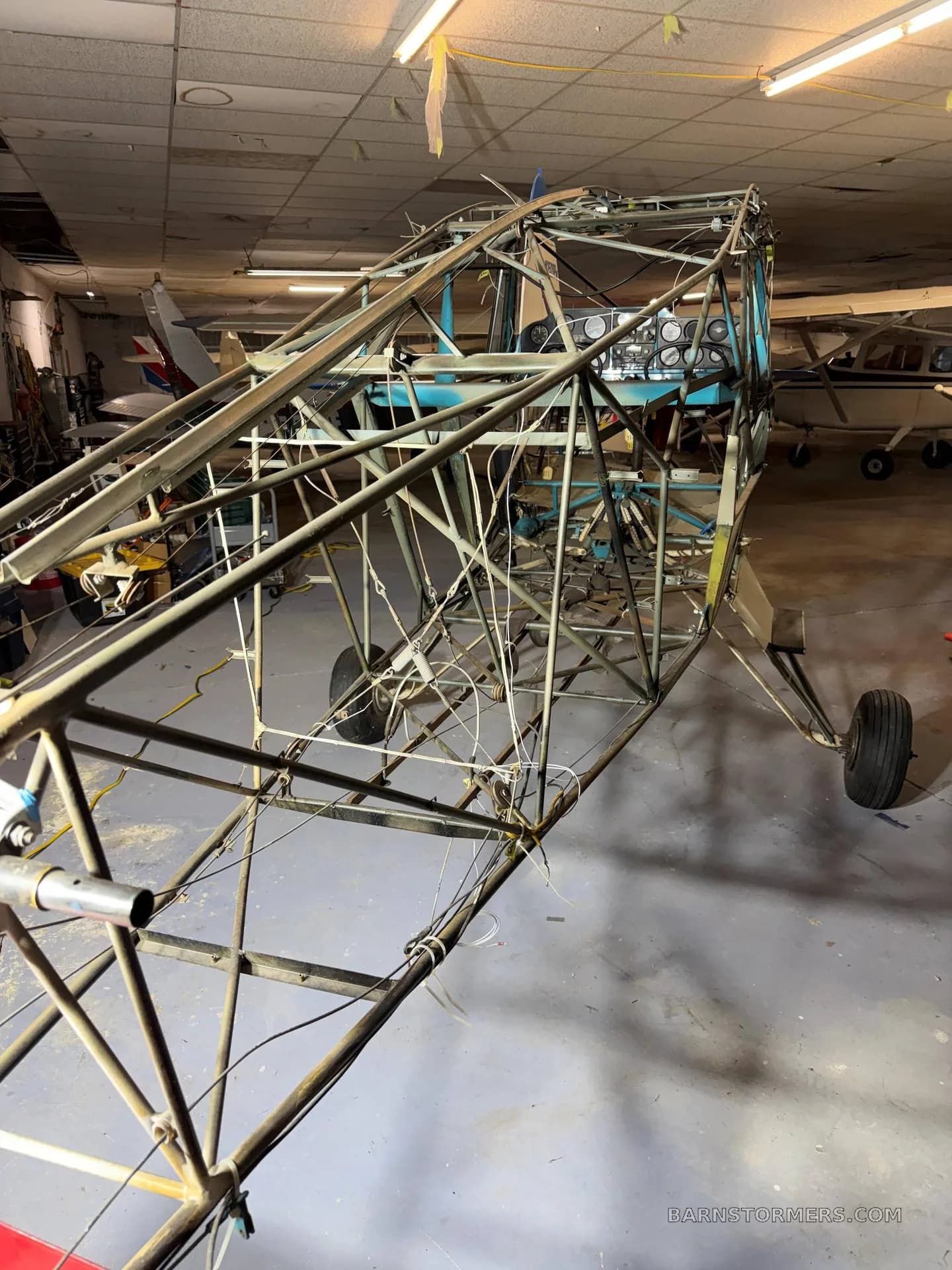 The bare metal frame of a Piper aircraft fuselage, with its cockpit panel visible, sits in a hangar.