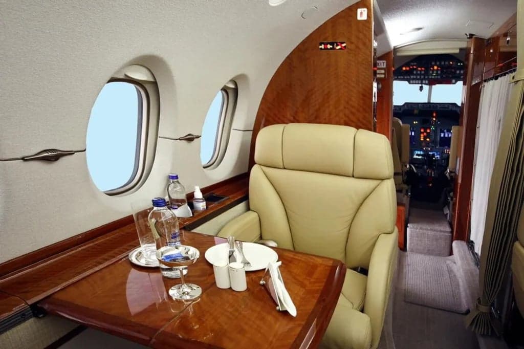 A view of the aircraft's interior with a tan leather seat, wooden table, and cockpit in the background.