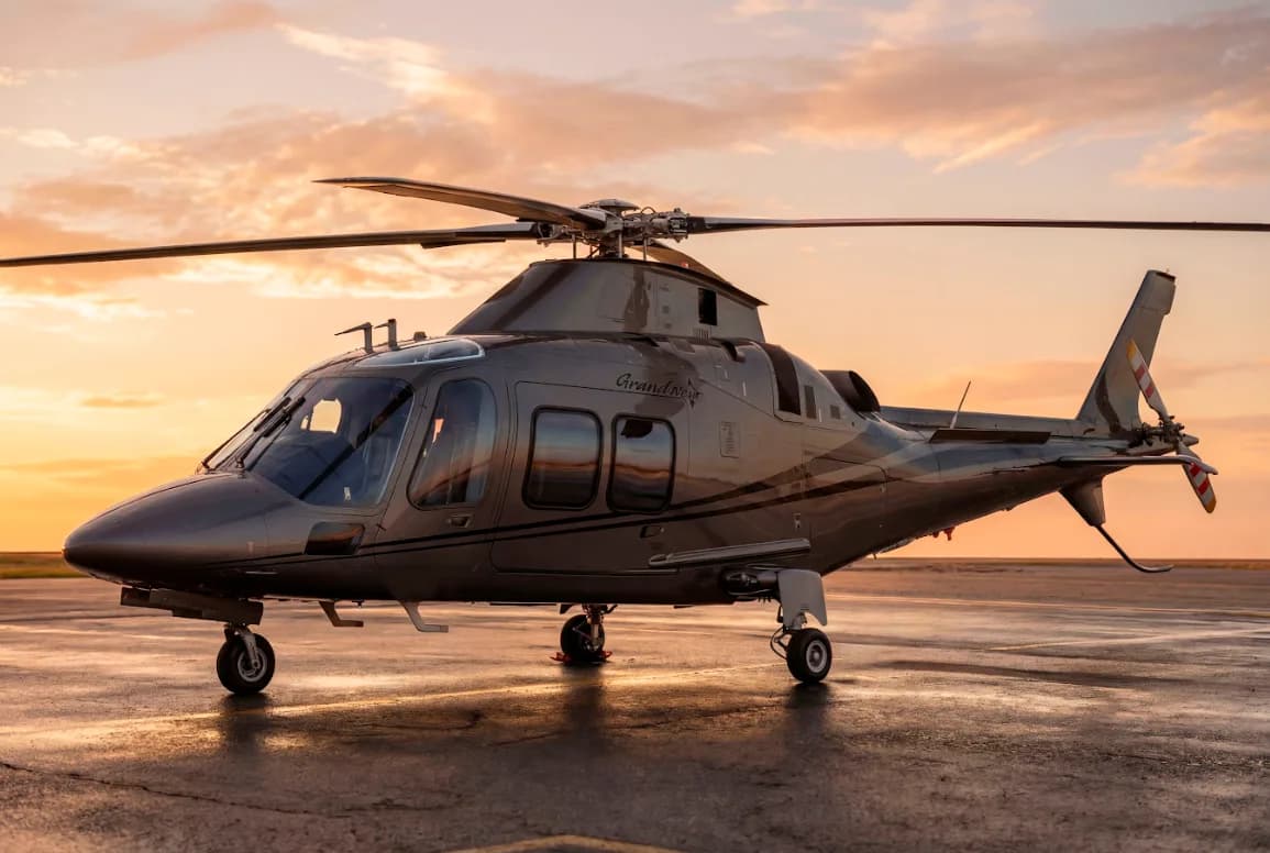 A grey AgustaWestland AW109 GrandNew helicopter sits on a wet tarmac at sunset.