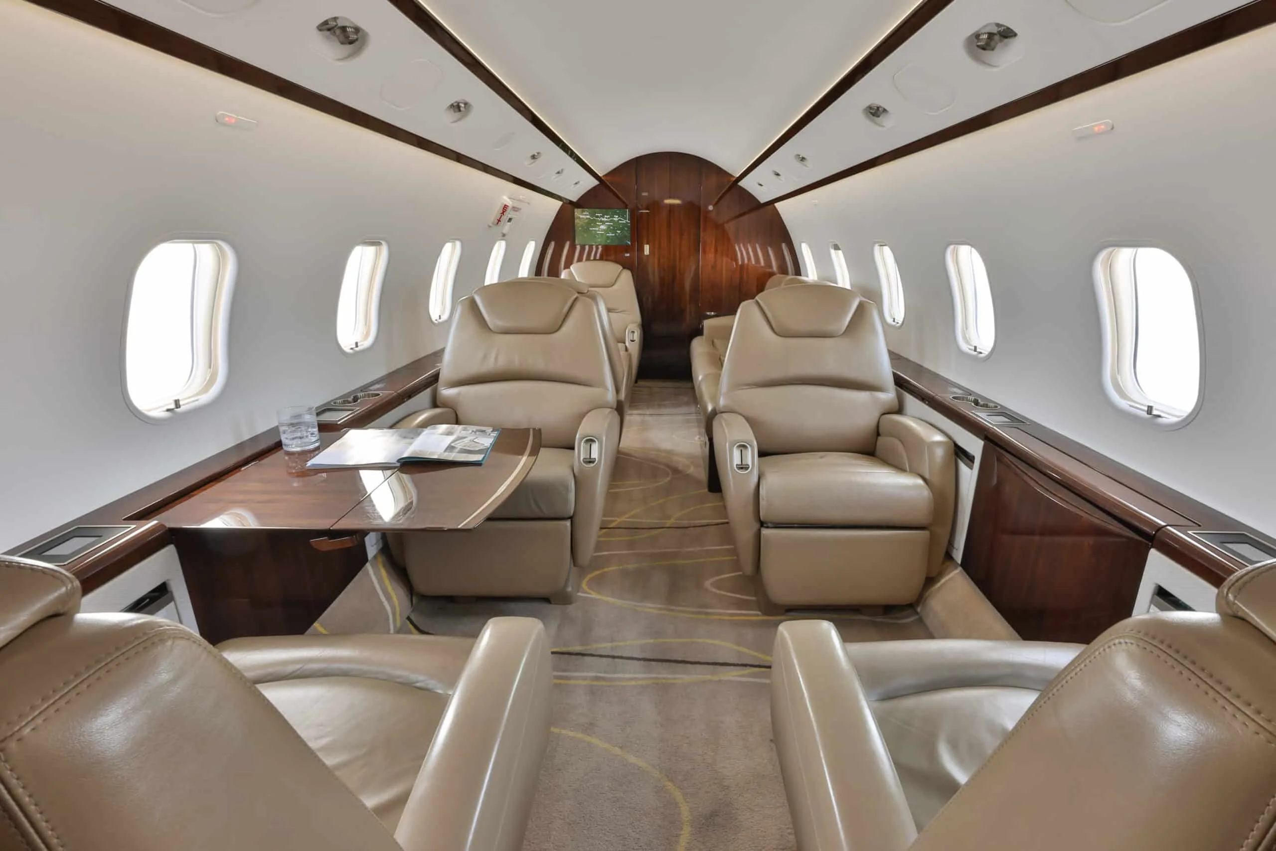 A wide view of the private jet cabin interior shows multiple beige leather club seats and dark wood accents.