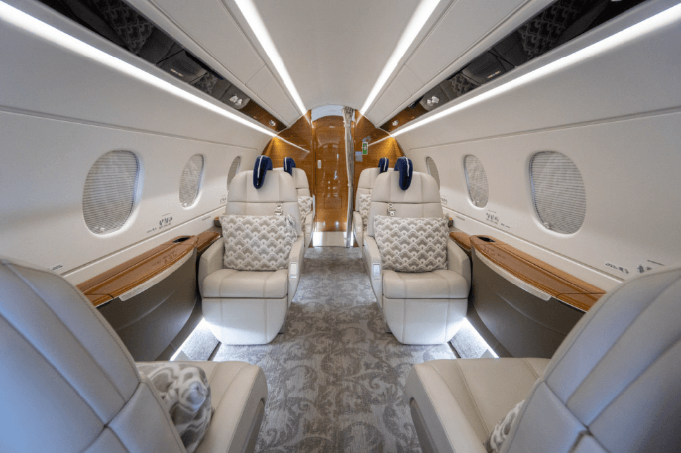 The elegant main cabin of a private jet, featuring light-colored leather seats with patterned pillows, wood trim, and overhead lighting.