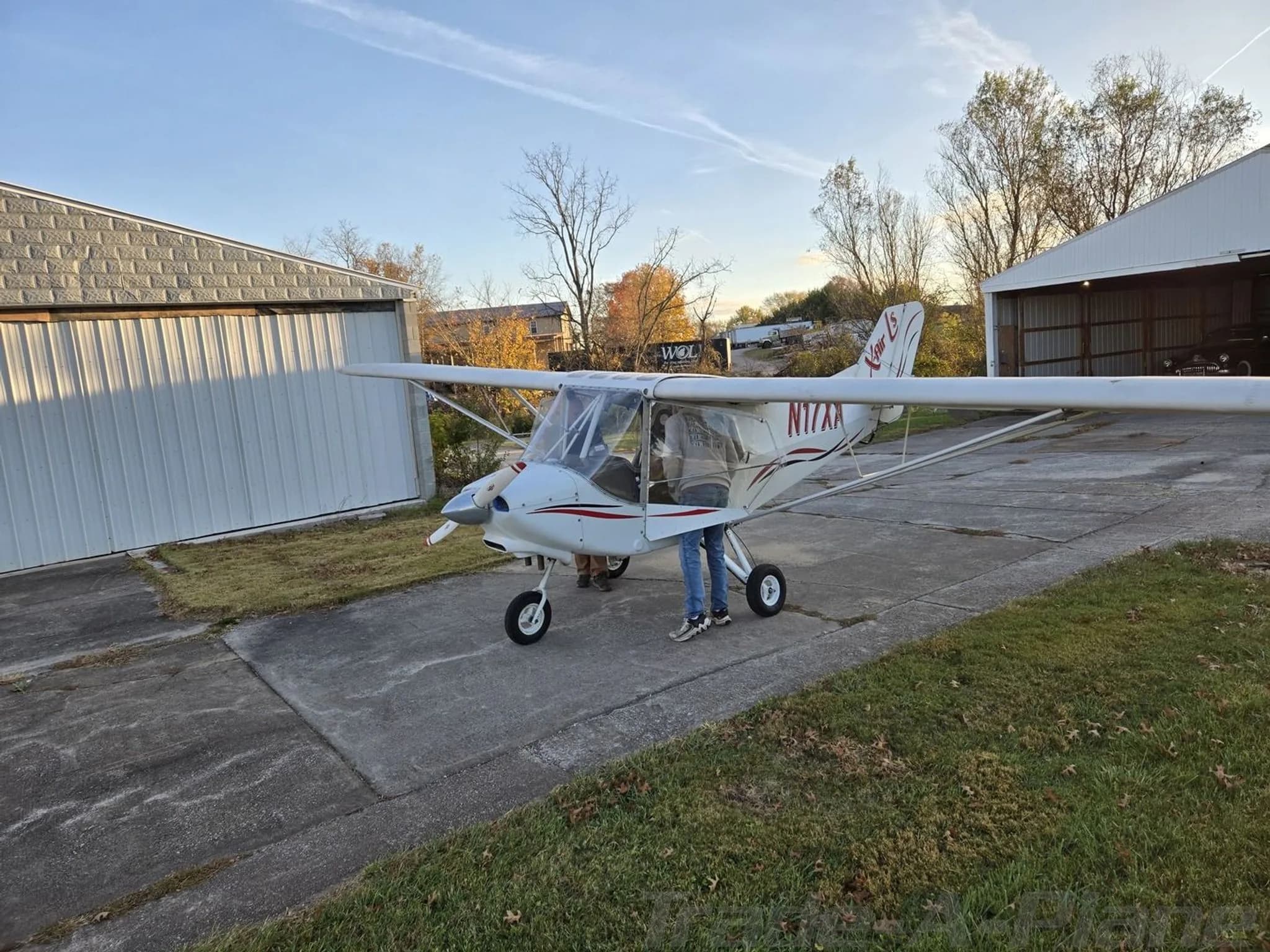 A white X-Air aircraft, registration N17XA, is parked on a concrete pad between two hangars.