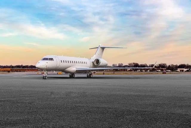 A white private jet, likely a Bombardier Global, parked on an airport tarmac under a colorful sky.