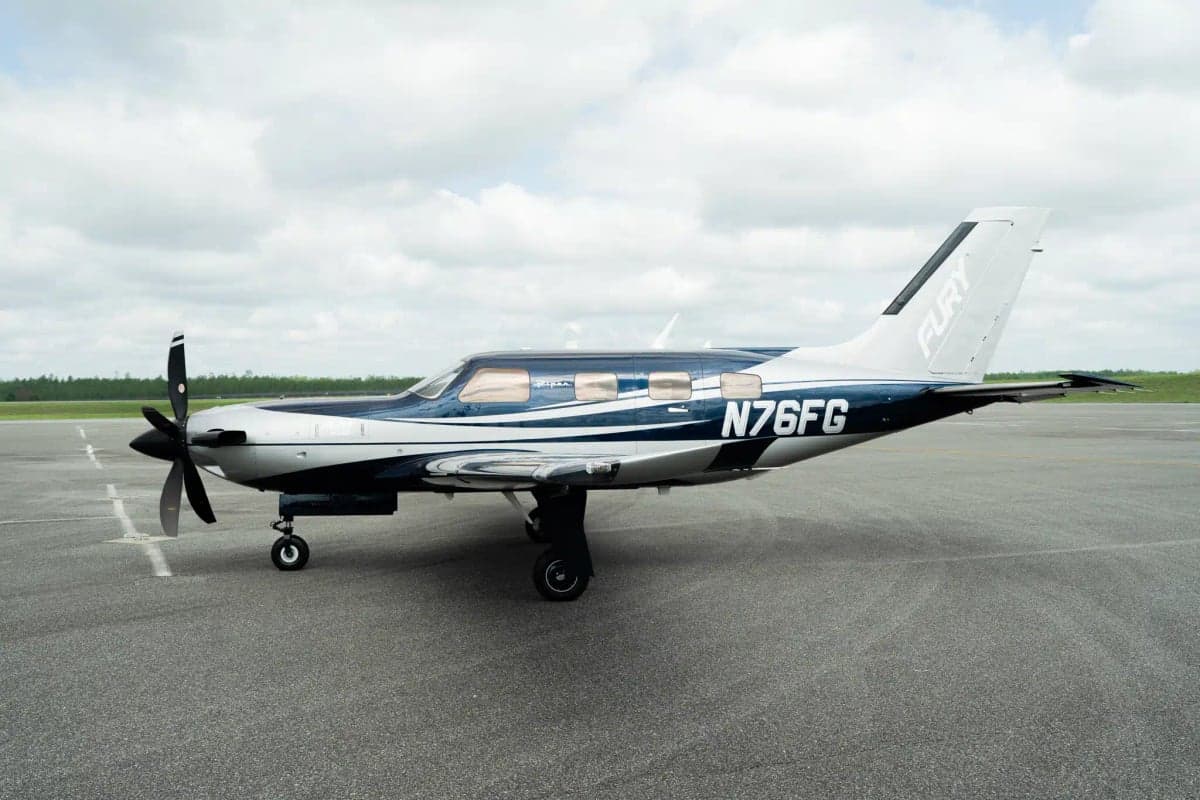 A Piper M700 Fury aircraft with a blue and silver paint scheme, registration N76FG, parked on an airfield under a cloudy sky.