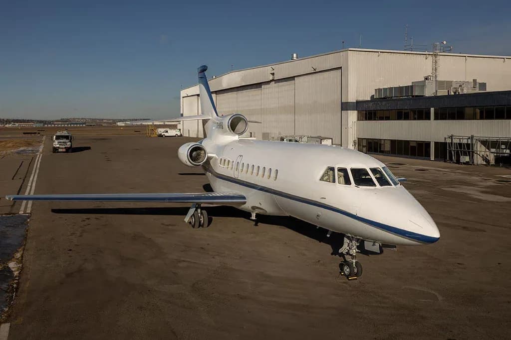 An aerial view of a white and blue Dassault Falcon 900EX business jet parked on an airfield next to hangars.