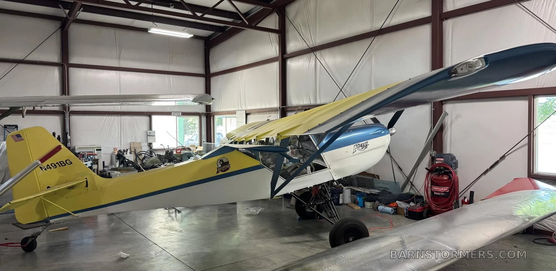 A yellow and blue Kitfox aircraft with its wing folded, parked in a hangar.