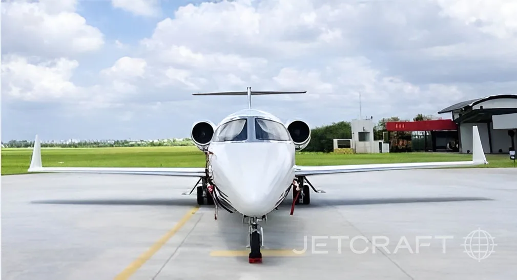 A white business jet with a black and grey stripe parked on the tarmac, viewed from the front.