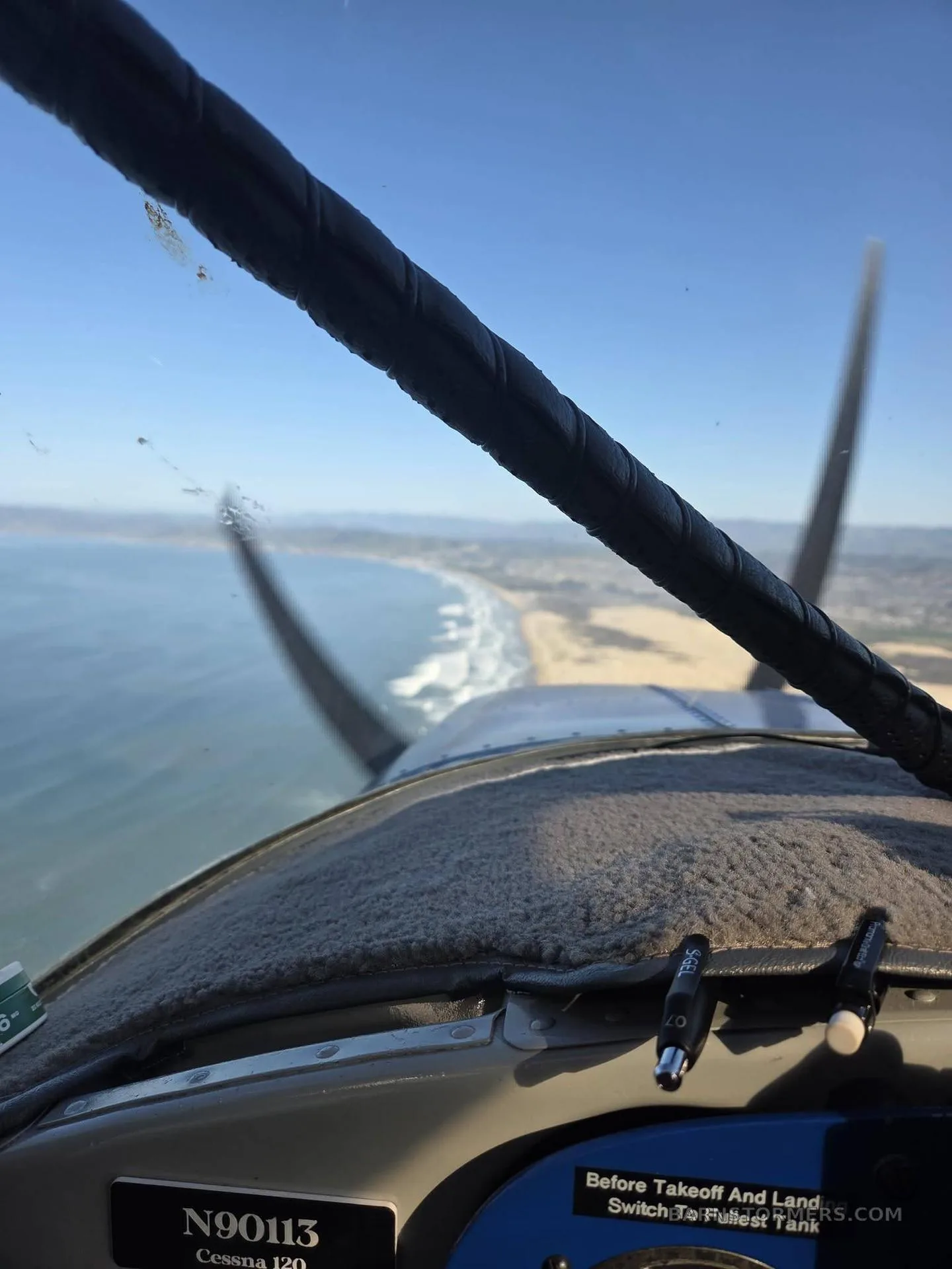 A view from the cockpit of a Cessna 120 flying along a sunny coastline with a beach and ocean.