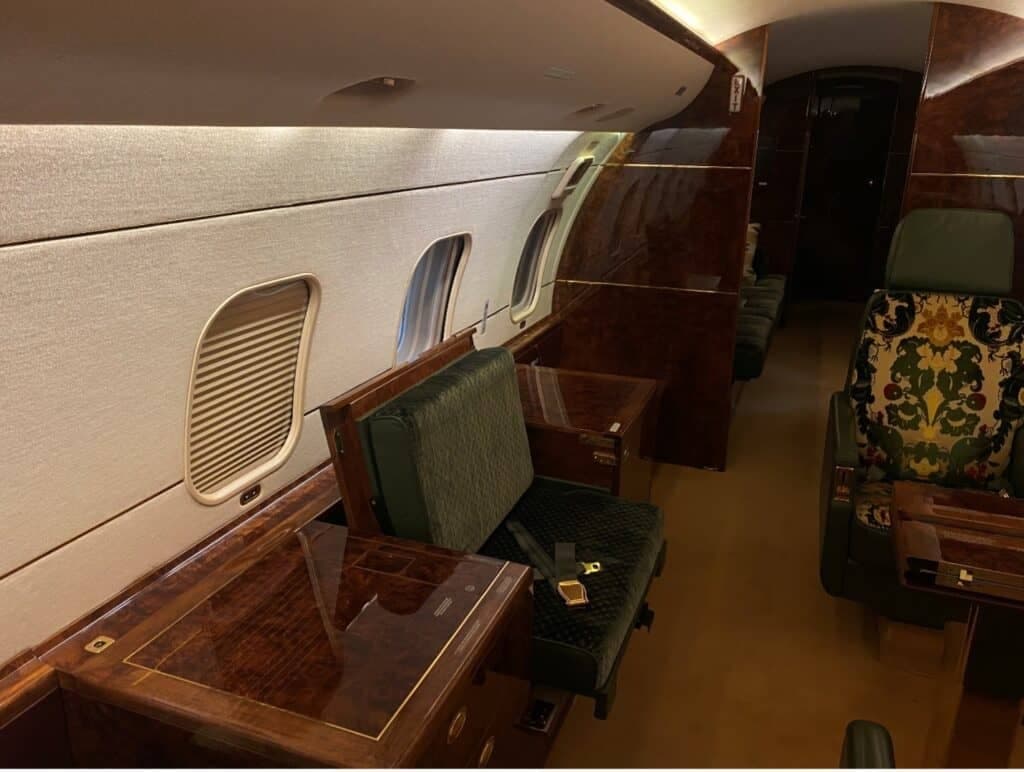 The luxurious interior cabin of a Bombardier Challenger 604, featuring dark wood paneling, green velvet seats, and patterned upholstery.