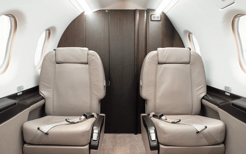 Two beige leather passenger seats in the cabin of a private aircraft, facing a dark wood partition.