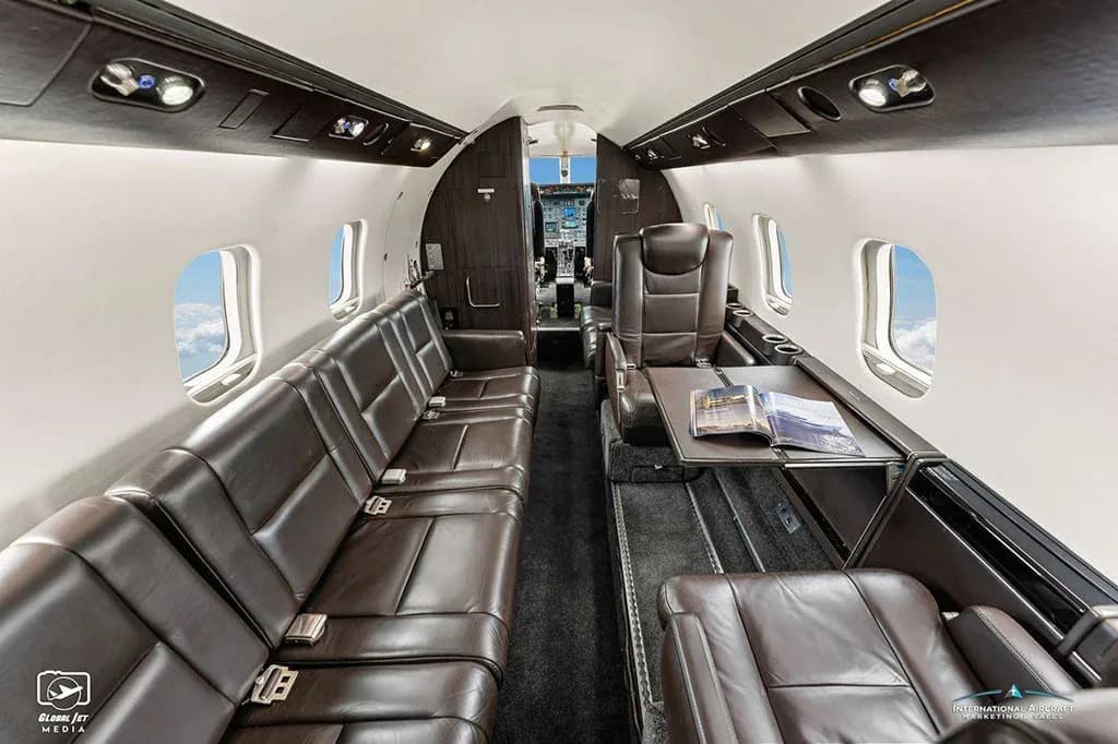 A wide view of the business jet's cabin, showing rows of brown leather seats and a table with a magazine.