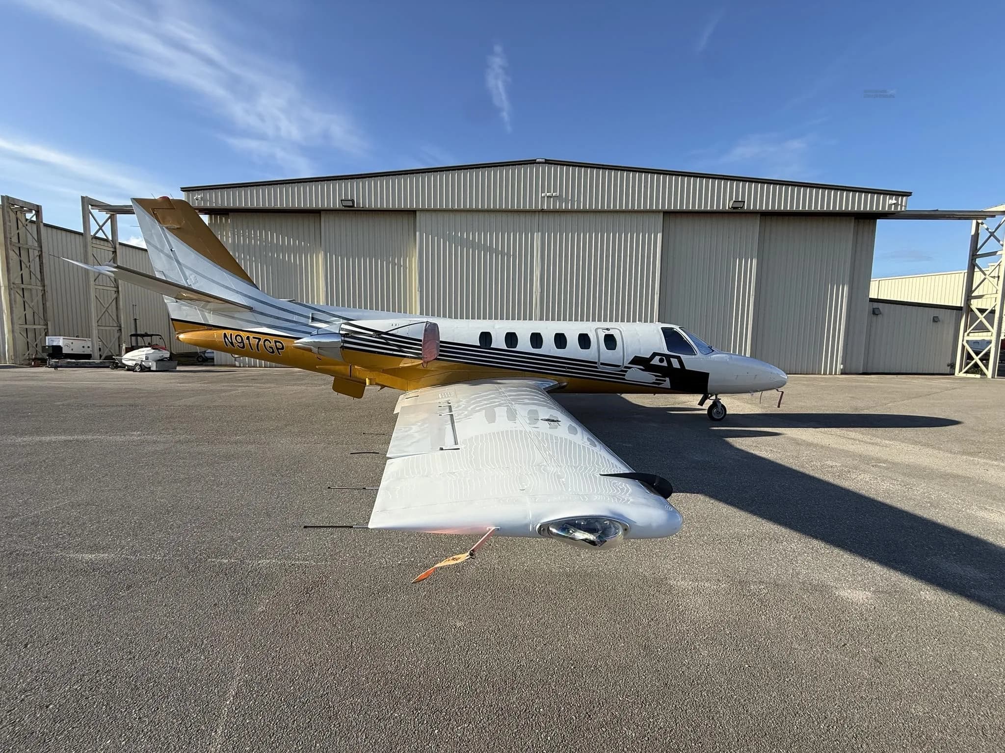 A Cessna Citation jet, N917GP, with white and gold paint scheme, parked on an airfield next to a hangar.