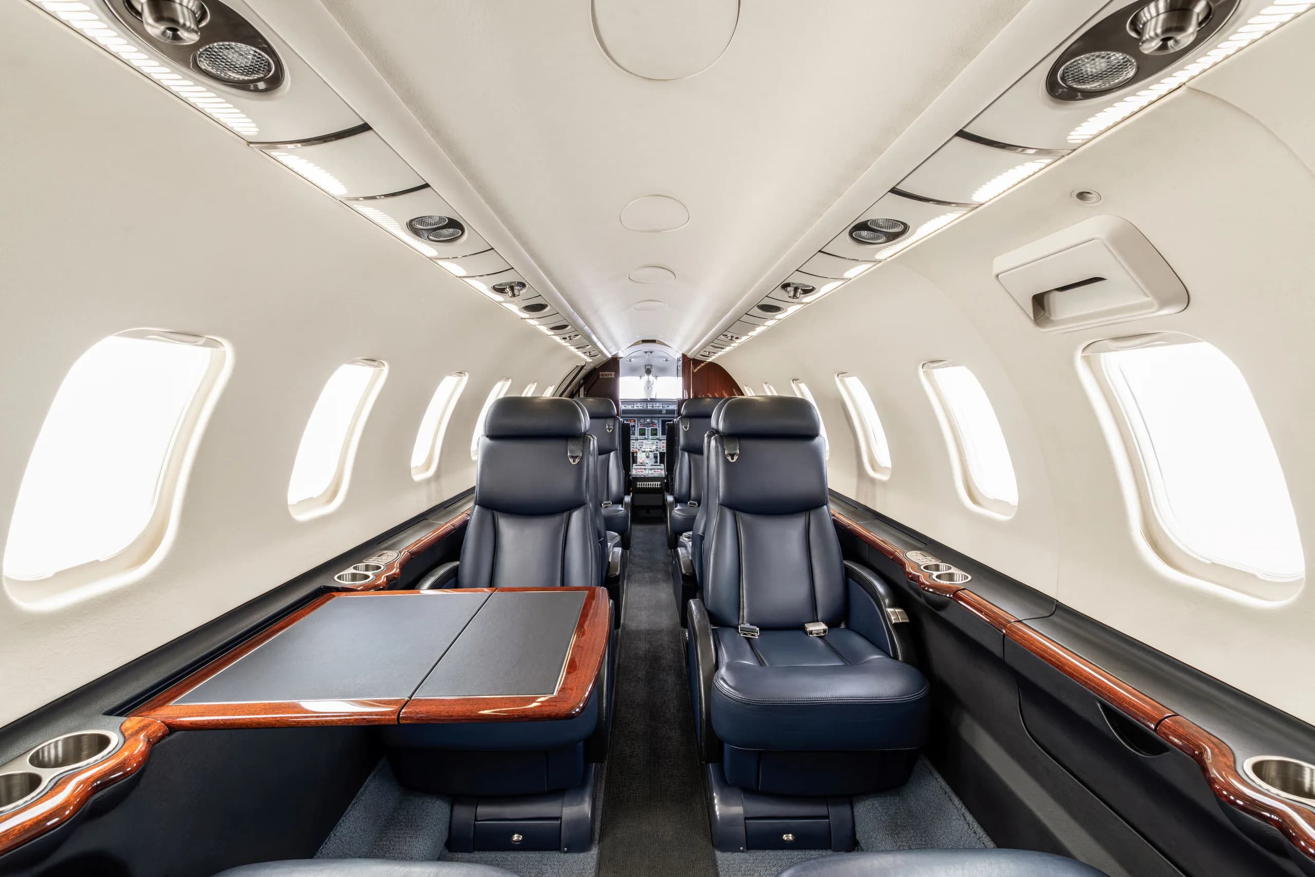 The interior cabin of a private jet, showing dark blue leather seats, a fold-out table, and light-colored walls with wood trim.