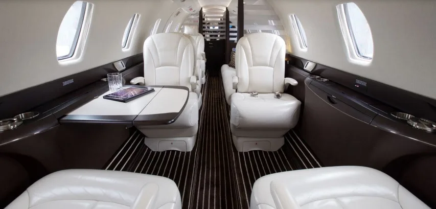 The elegant cabin of a private jet features white leather seats, a table with a tablet, and dark striped flooring.