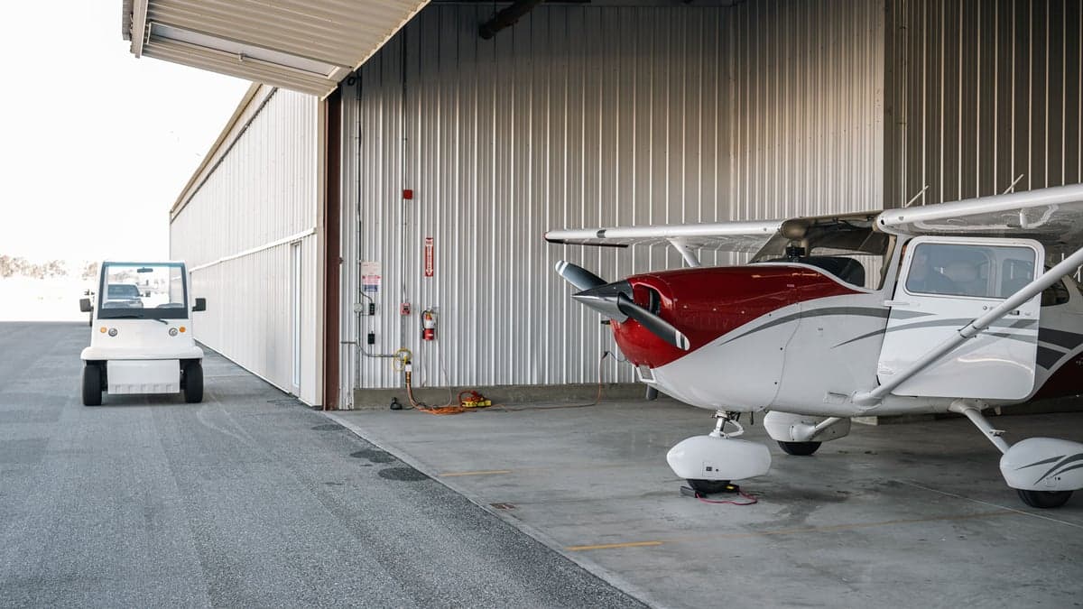 A white and red single-engine propeller aircraft partially parked in a hangar with a white utility vehicle outside.