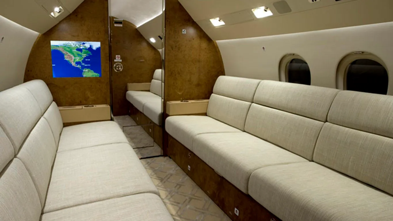 A section of the aircraft cabin featuring two long beige sofas facing each other, with wood paneling and a display screen.