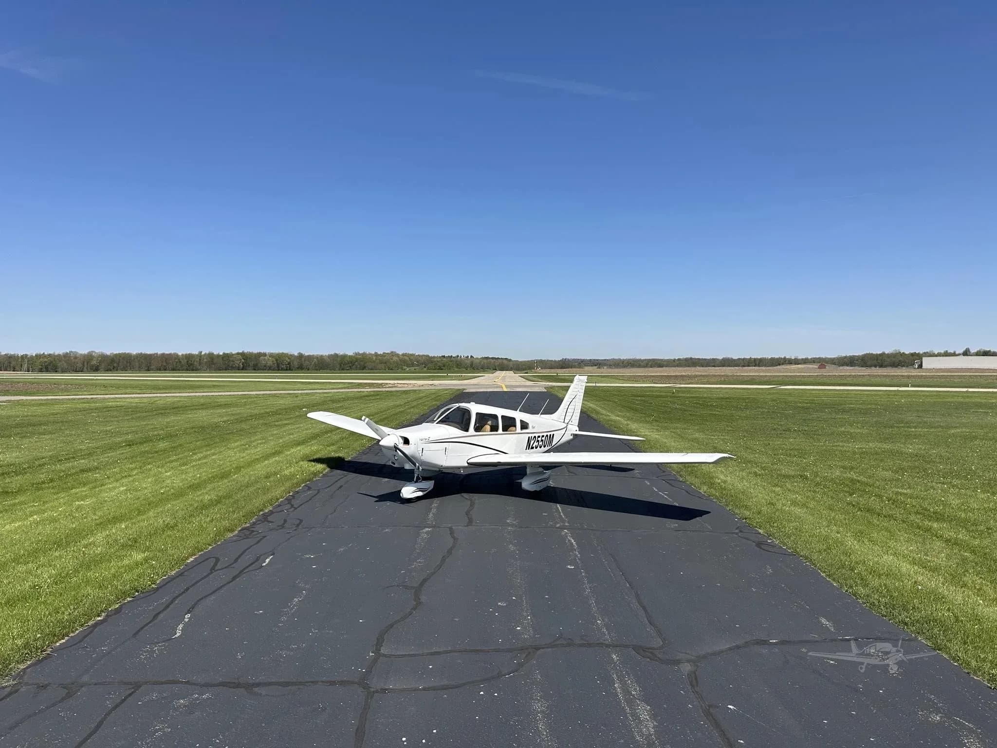 A white Piper Cherokee aircraft, N2550M, parked on an asphalt runway, with a clear blue sky and distant trees.