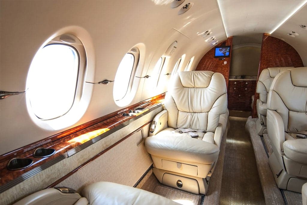 A view inside the business jet cabin, highlighting cream leather seating, polished wood accents, and bright windows.