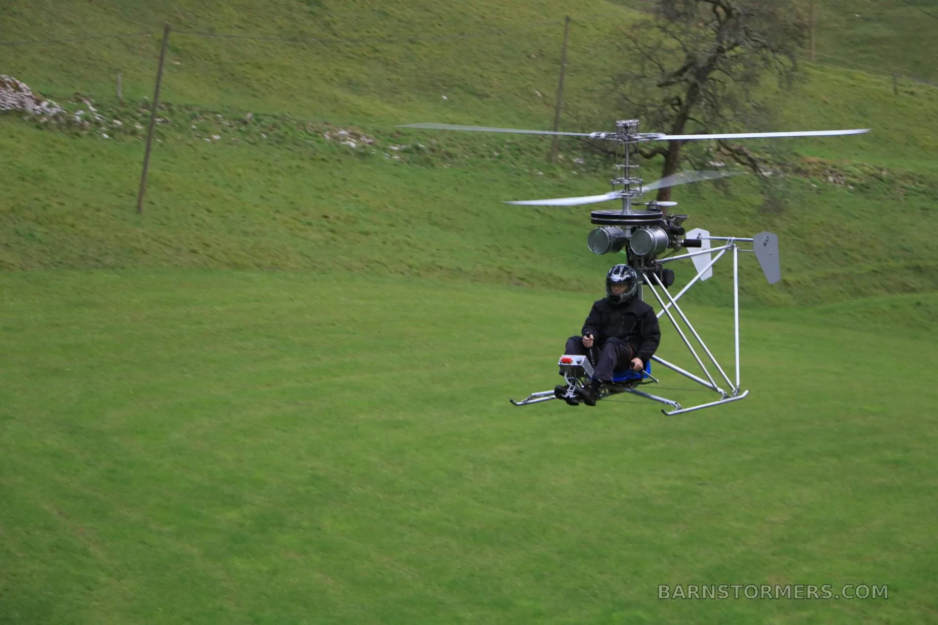 A single-seat ultralight helicopter with a pilot in black gear hovers over a green grassy field.